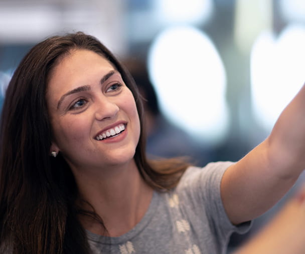 Woman with long brown hair and wearing a gray shirt points to something on a board while collaborating with another person