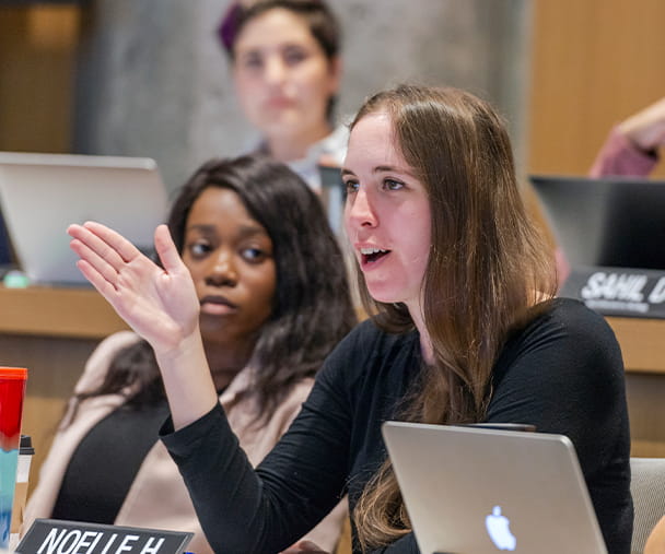 A Kellogg School of Management student actively participates in a classroom discussion, gesturing expressively. She is seated among peers, some of whom have laptops open in front of them. The environment reflects a dynamic and collaborative learning atmosphere.